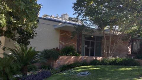 One-story building with flat roof, a stucco wall with brick panel facade and metal panels lining the top, glass front doors, and a sloping lawn with grass and trees
