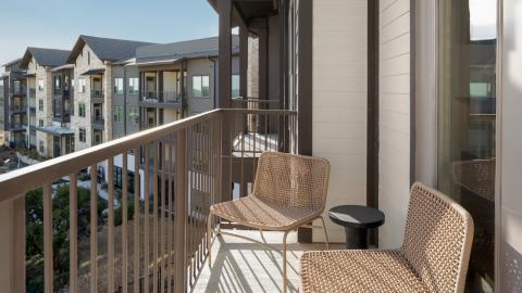 Photo or rendering or mix of an apartment balcony with two chairs and a small table in front of a sliding glass door looking out over a large, mid-rise apartment complex.